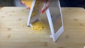 Grating cheddar cheese on a wooden board with a white grater.